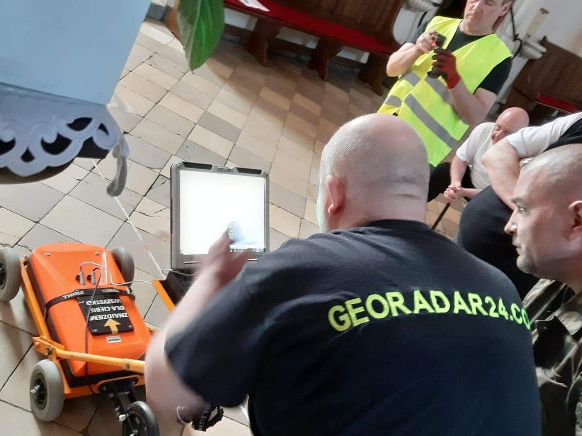 Ekipa georadar24.com podczas badania georadarem we wnętrzu kościoła — pomarańczowy wózek georadaru i laptop z podglądem wyniku, technik w koszulce z napisem Georadar24.com omawia pomiar z obecnymi.